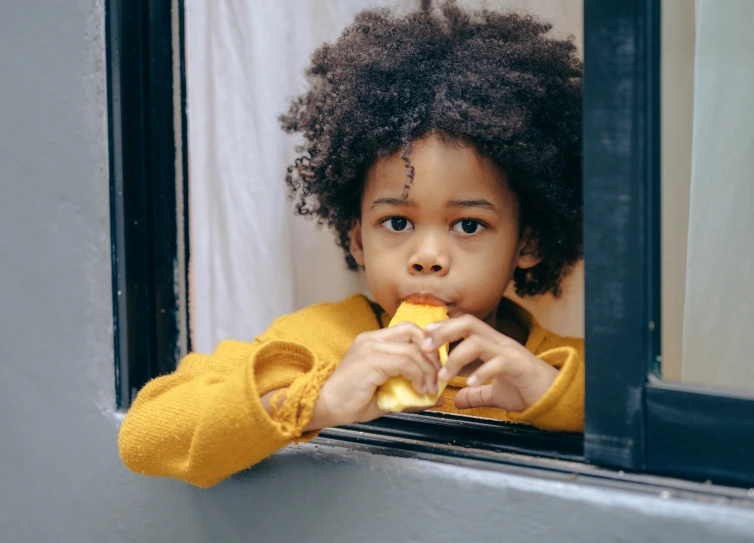 a close up of a child eating a banana, by Winona Nelson, pexels contest winner, near a window window, afro comb, yellow awning, thoughtful pose