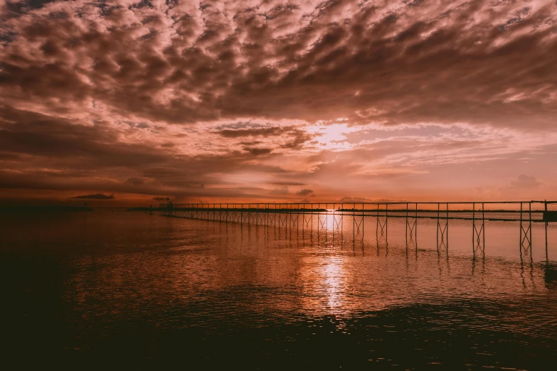 a boat sitting on top of a body of water under a cloudy sky, it is sunset, bridge over the water, unsplash photo contest winner, faded red colors