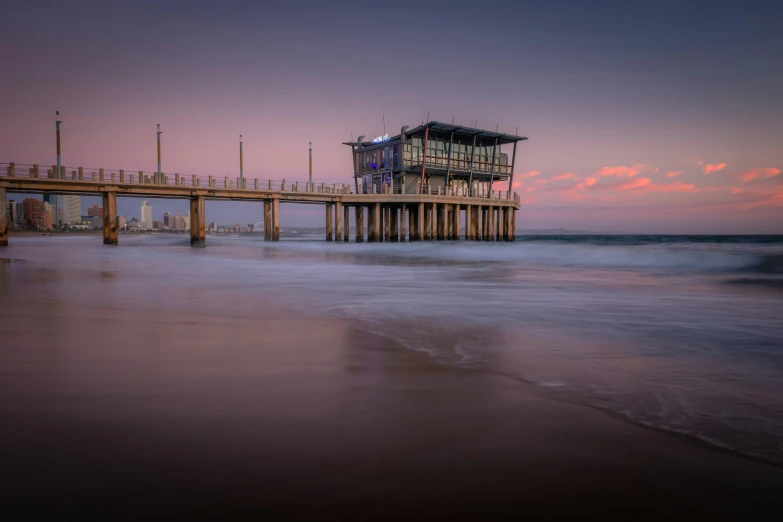 a pier sitting on top of a sandy beach next to the ocean, by Lee Loughridge, unsplash contest winner, renaissance, blue hour, south african coast, pink sunset hue, thumbnail