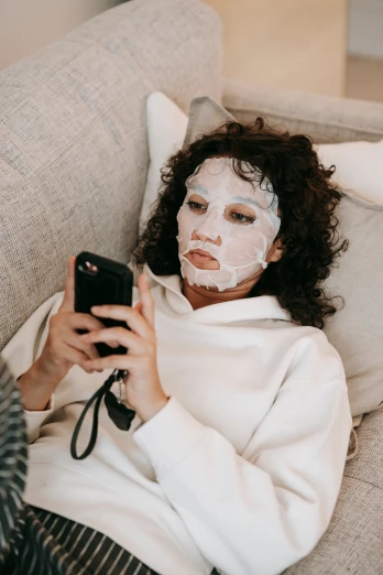 a woman laying on a couch with a face mask on, by Julia Pishtar, trending on pexels, happening, goddess checking her phone, white face makeup, jordan peele's face, brunette
