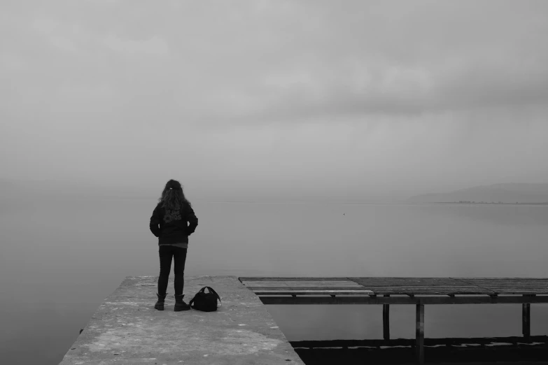 a black and white photo of a person standing on a dock, by Emma Andijewska, teenage girl, sad sky, isolated, girl standing