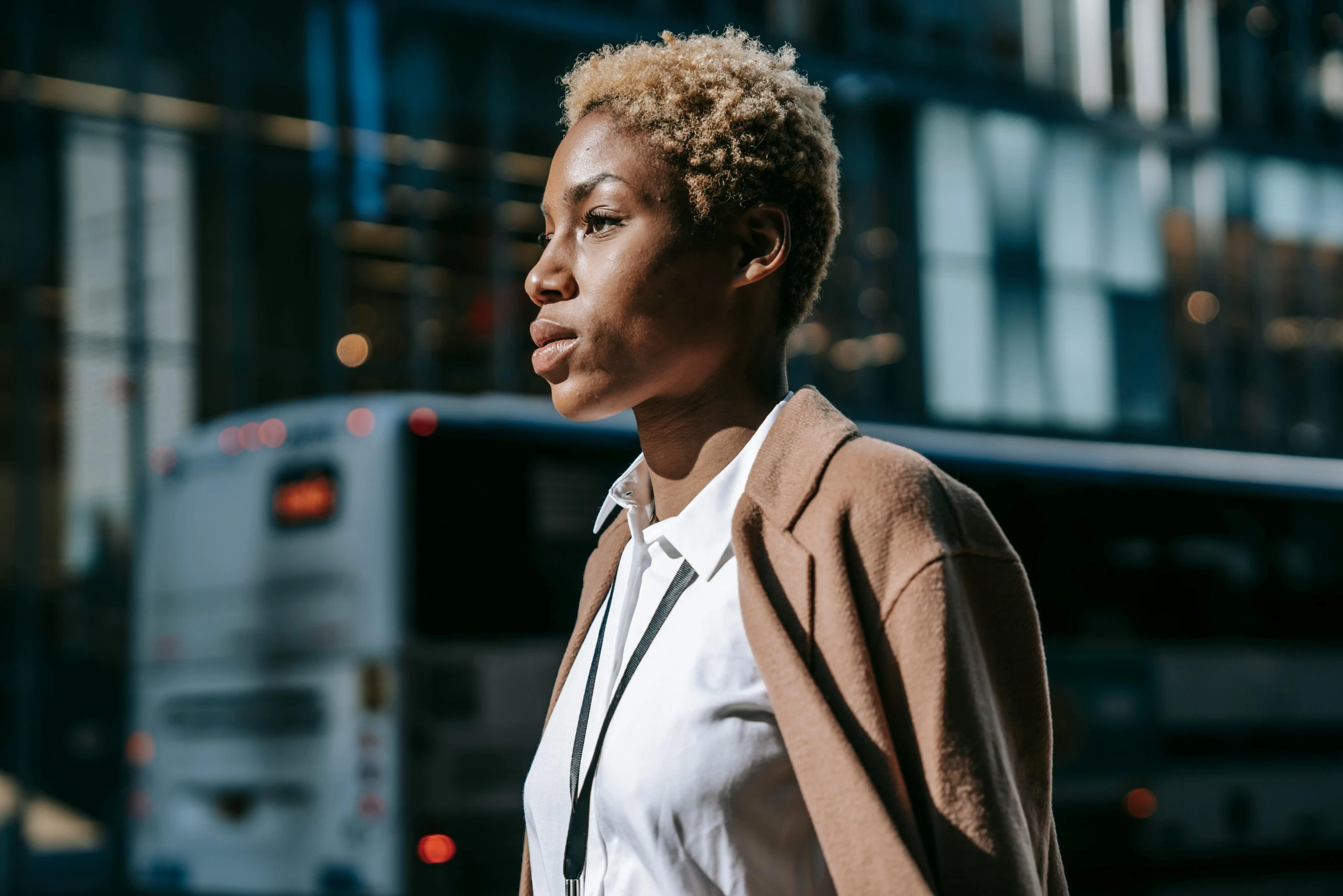 a woman walking down a street with a bus in the background, by Carey Morris, pexels contest winner, afrofuturism, short blonde afro, thoughtful expression, evening sunlight, thumbnail