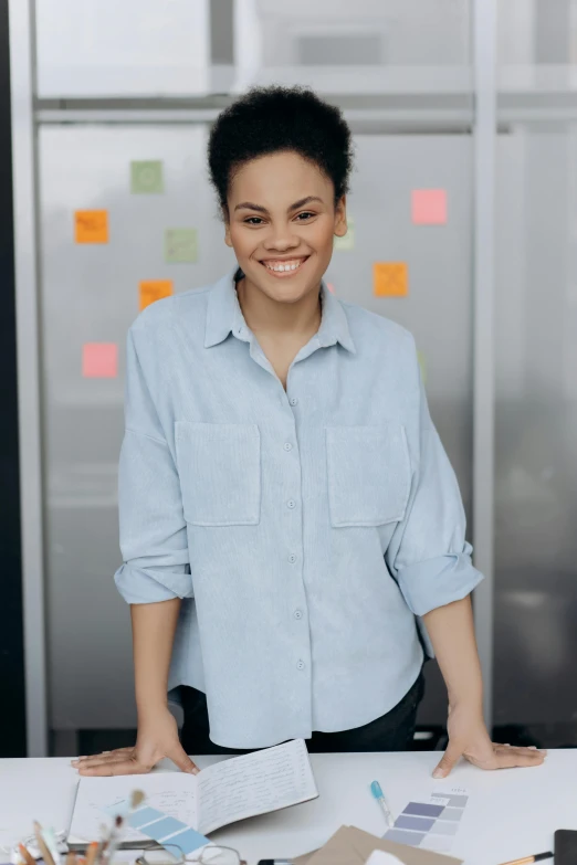 a woman standing in front of a table with papers, wearing a light blue shirt, smiling confidently, 2019 trending photo, thumbnail