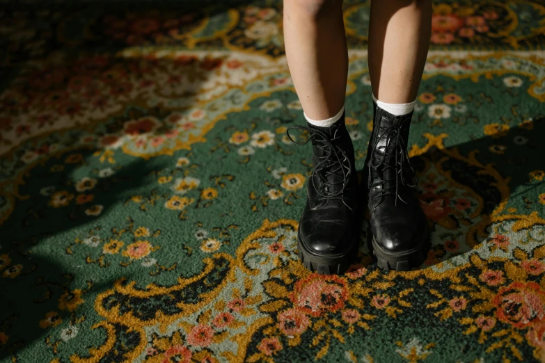 a close up of a person's shoes on a rug, an album cover, inspired by Elsa Bleda, trending on pexels, visual art, girl wearing uniform, ornate dark green clothing, wearing black boots, persian carpet
