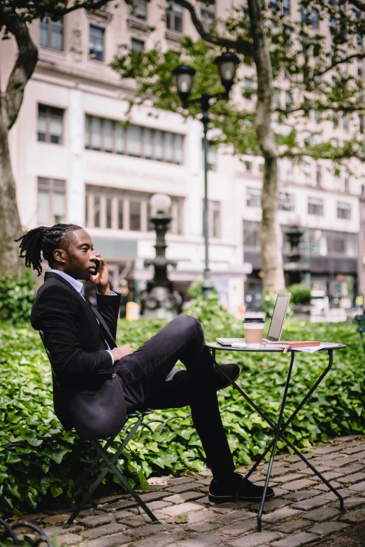 a man sitting on a bench talking on a cell phone, a black man with long curly hair, with a business suit on, lush brooklyn urban landscaping, sitting on a mocha-colored table