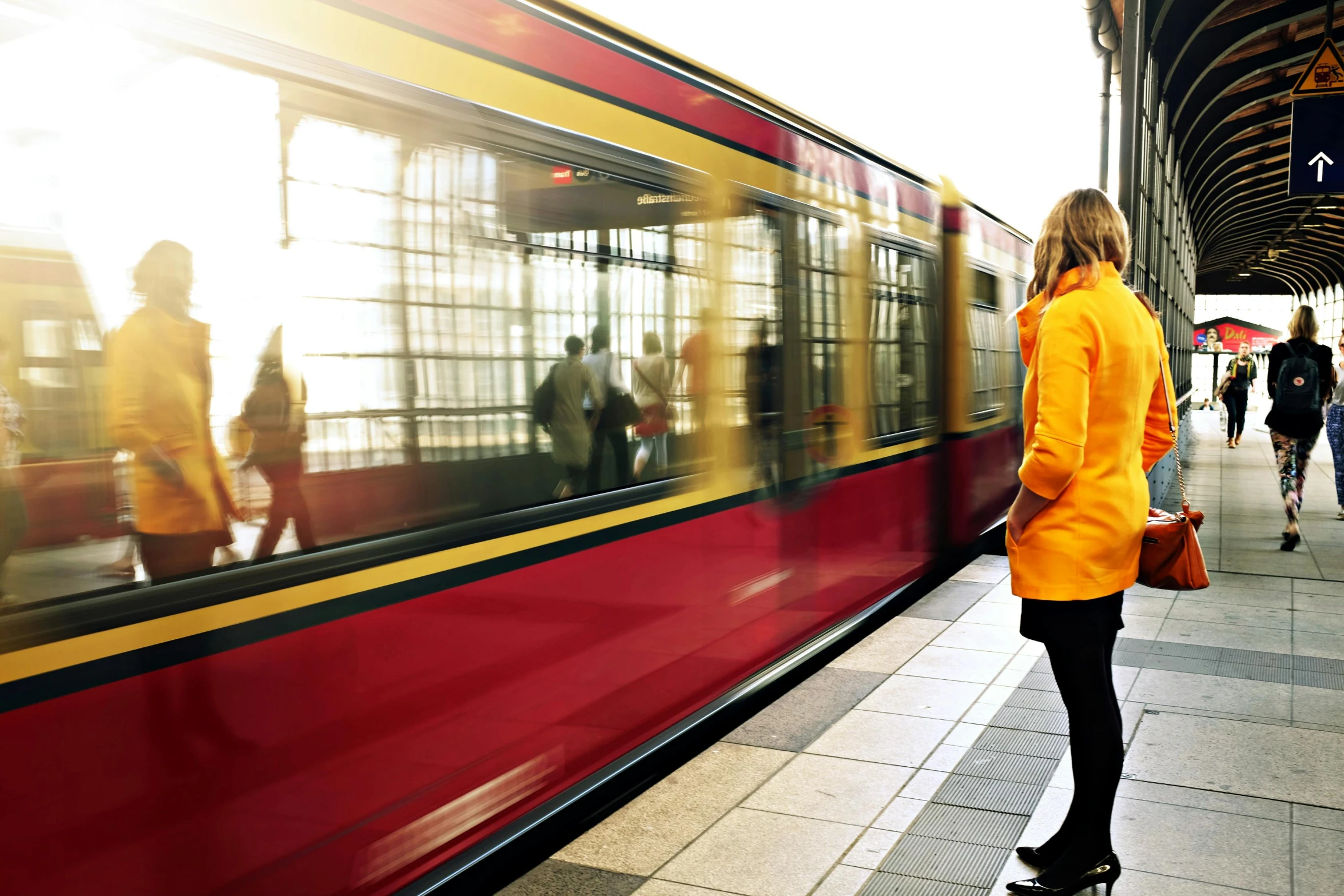 a woman waiting for a train at a train station, by Thomas Häfner, unsplash, happening, wearing red and yellow clothes, trams ) ) ), yellow and black, charging through city