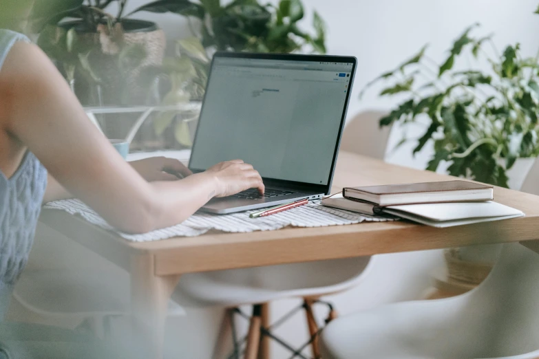 a woman sitting at a table using a laptop computer, trending on pexels, next to a plant, wooden desks with books, no - text no - logo, over-the-shoulder shot