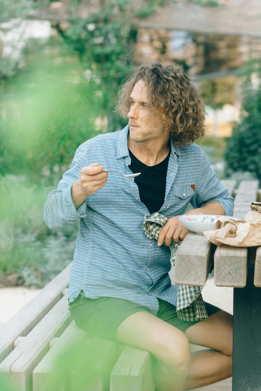 a man sitting on top of a wooden bench, eating outside, with long curly hair, wearing a linen shirt, style of seb mckinnon