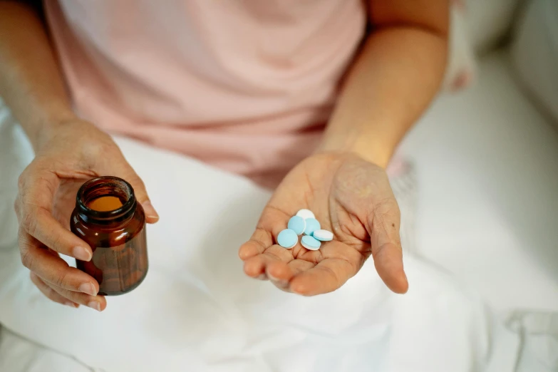 a woman sitting on a bed holding a bottle of pills, by Rachel Reckitt, pexels, antipodeans, blue robe, candy treatments, pictured from the shoulders up, pixivs and