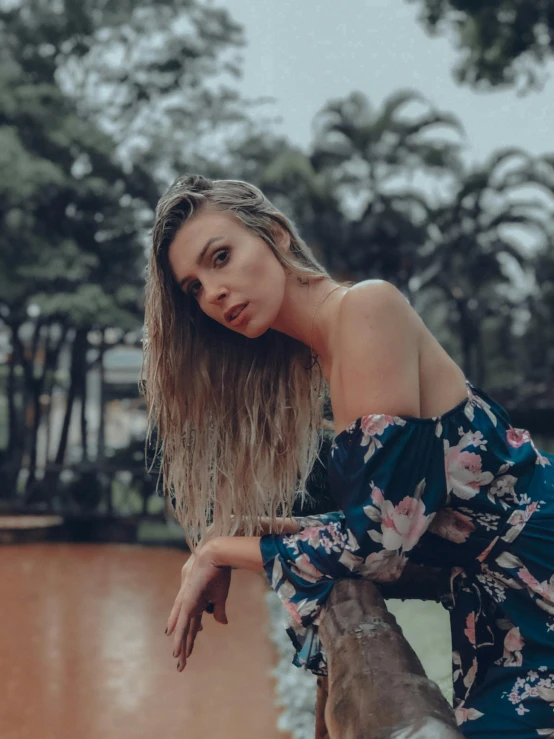 a woman sitting on a fence next to a body of water, a picture, inspired by Elsa Bleda, pexels contest winner, aestheticism, sexy look at the camera, in the jungle. bloom, profile image, floral clothes