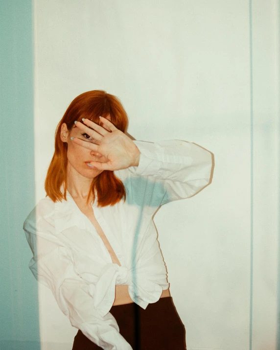 a woman standing in front of a mirror covering her eyes, an album cover, inspired by Elsa Bleda, trending on pexels, aestheticism, ginger hair, wearing a white button up shirt, hand gesture, non binary model