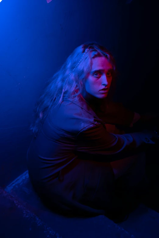 a man sitting on the ground in a dark room, an album cover, inspired by Nan Goldin, renaissance, long blonde hair and blue eyes, timothee chalamet, concert photo, menacing look