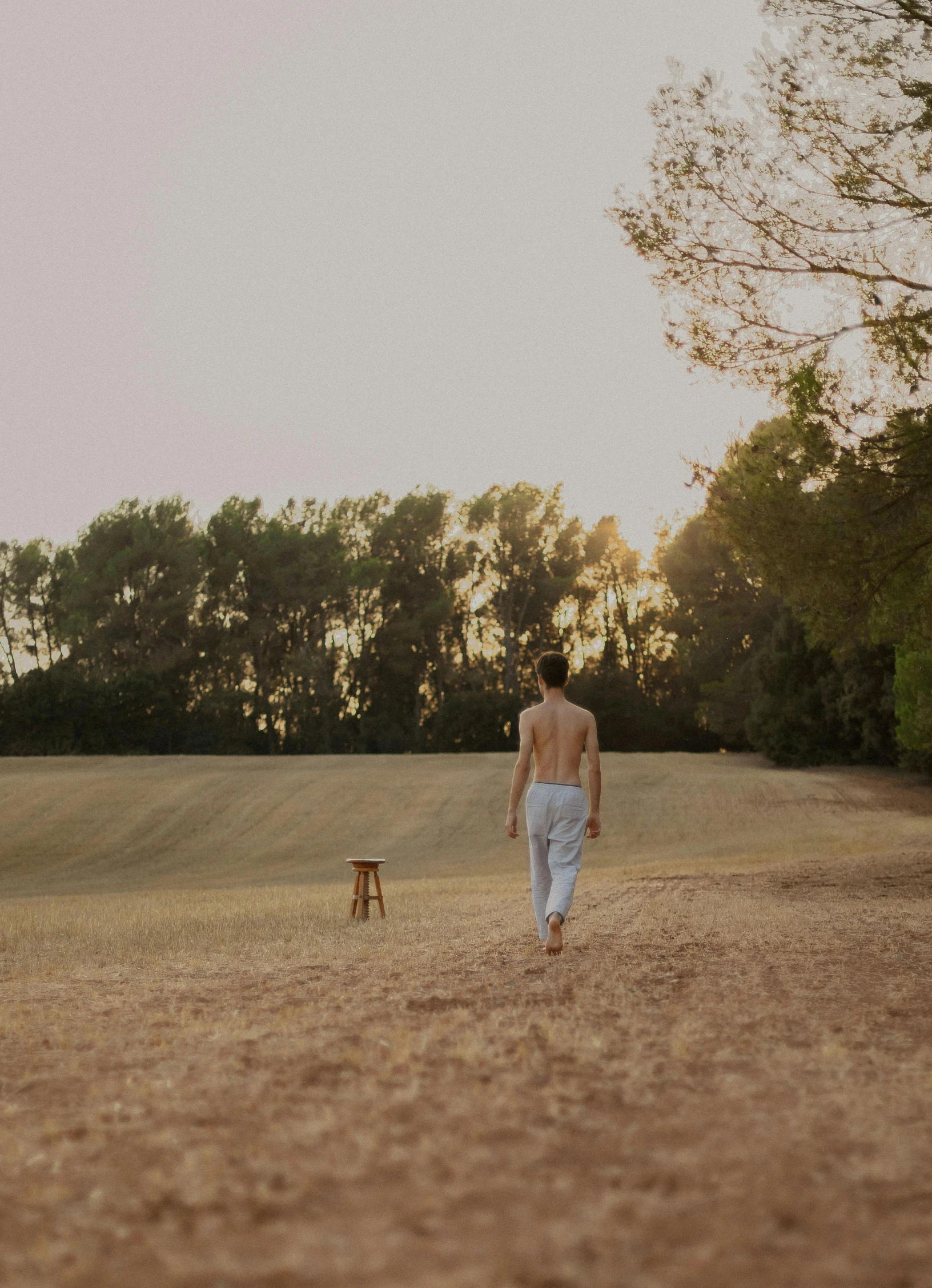 a man walking away from a chair in a field, a picture, unsplash, happening, in spain, bare shoulders, soft glow, on the sand