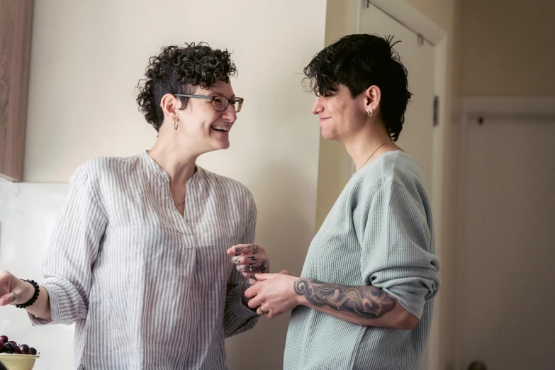 two women standing next to each other in a kitchen, a portrait, by Liza Donnelly, pexels, dark short curly hair smiling, tattooed, smiling at each other, promotional image