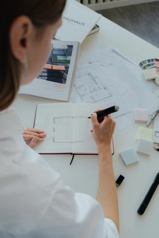 a woman sitting at a table working on a project, a drawing, trending on unsplash, white architecture, rounded corners, holding notebook, low quality photo