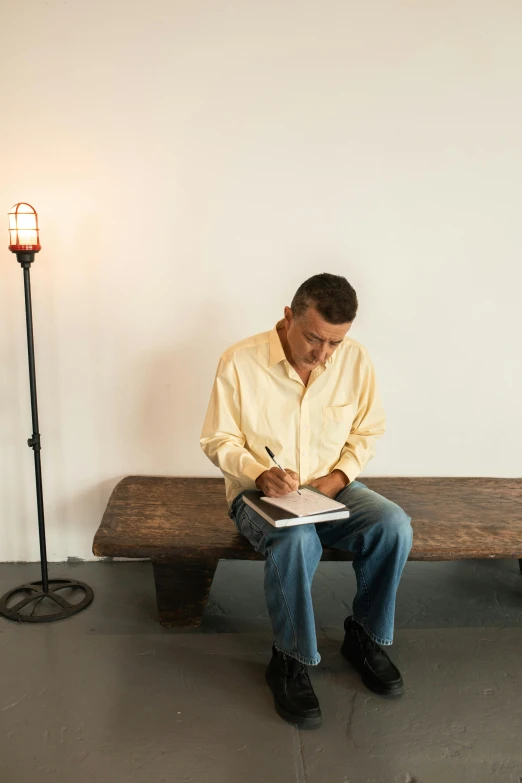 a man sitting on top of a wooden bench, writing in journal, lights off, brazilian ronaldo, in a gallery setting