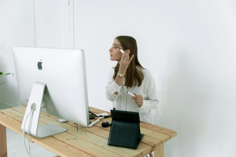 a woman standing in front of a computer on a desk, by Julia Pishtar, trending on pexels, her face is coated in a whitish, putting makeup on, minimalistic, people at work