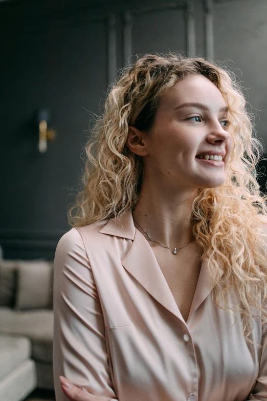 a woman standing in a living room next to a couch, a character portrait, by Adam Marczyński, trending on pexels, renaissance, curly blonde hair, earing a shirt laughing, closeup headshot, elegant profile pose