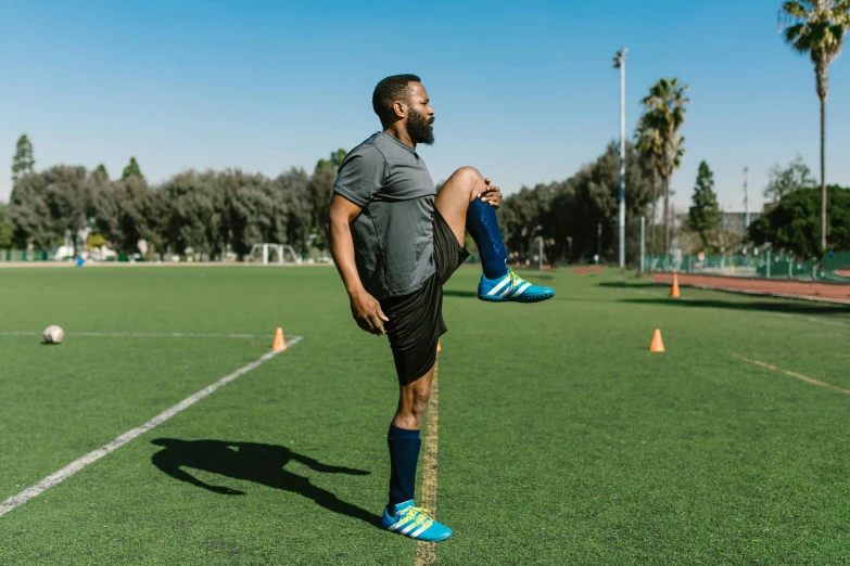 a man standing on top of a soccer field, stretching her legs on the grass, origin jumpworks, gray shorts and black socks, riyahd cassiem