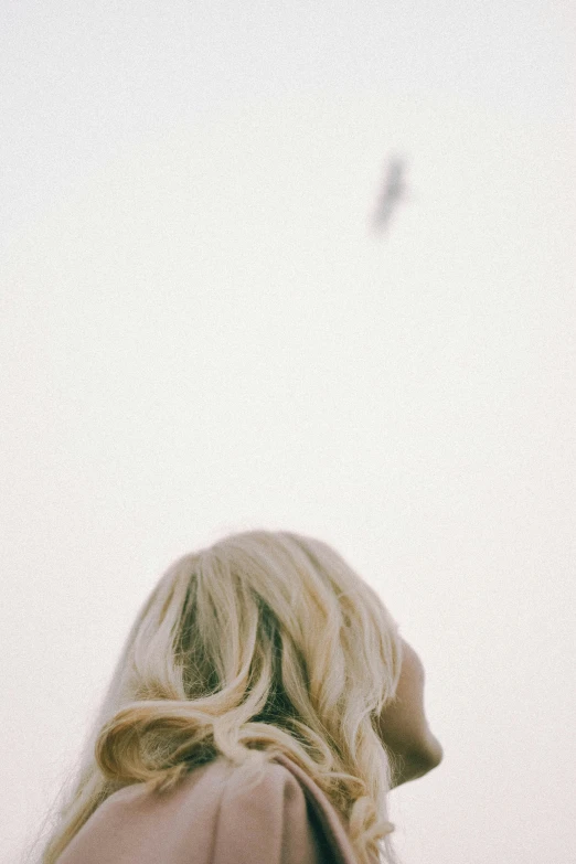 a woman standing in front of a bird flying in the sky, inspired by Ren Hang, unsplash, postminimalism, close up of a blonde woman, pale beige sky, back of the hair, distant - mid - shot