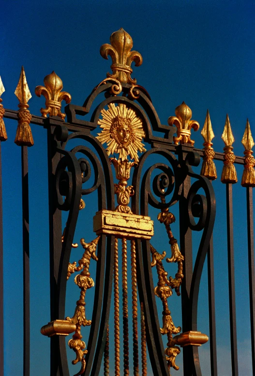a close up of a metal fence with a clock on it, an album cover, inspired by Prince Hoare, baroque, ornate gold crown, blue sky, golden sun, interior of buckingham palace