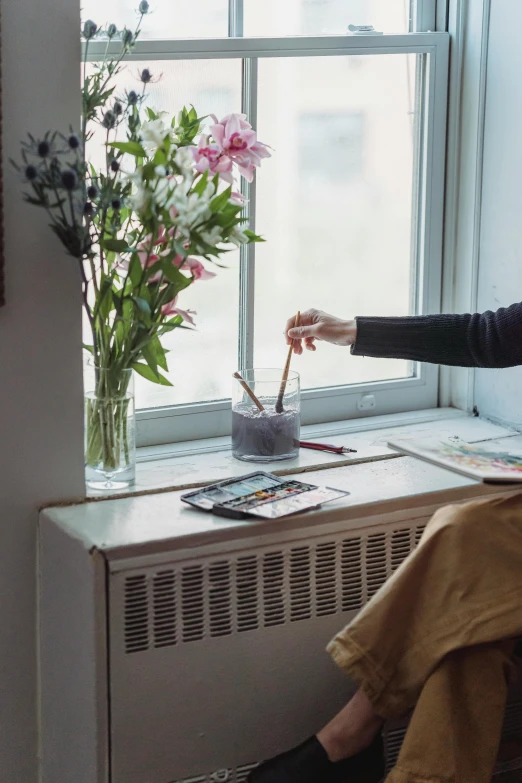 a woman sitting on a window sill next to a vase of flowers, a photorealistic painting, inspired by Elsa Bleda, pexels contest winner, holding a paintbrush in his hand, apartment of an art student, swiping brushwork, color photograph