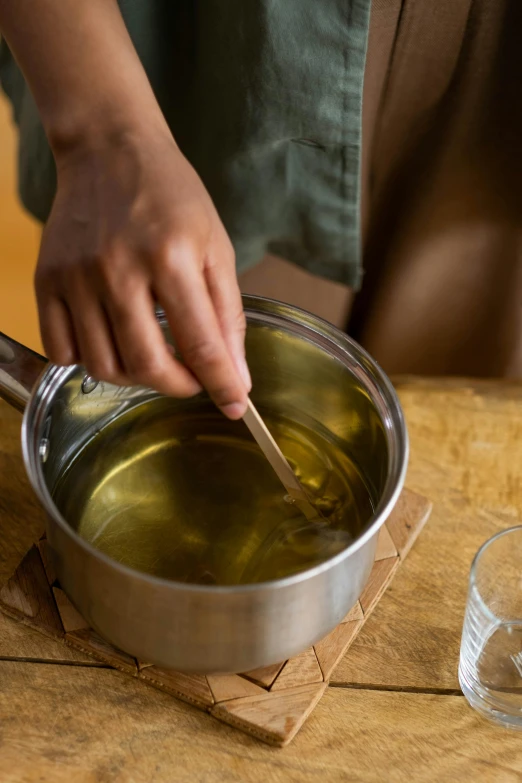 a person stirring olive oil in a saucepan, by Nina Hamnett, process art, candle wax, promo image, at home, clear liquid