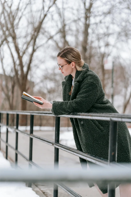 a woman leaning on a railing reading a book, inspired by Louisa Matthíasdóttir, pexels contest winner, wearing hunter coat, reading engineering book, cold temperature, wearing green jacket