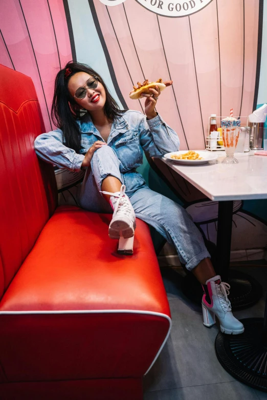 a woman sitting in a booth eating a slice of pizza, an album cover, by Bernie D’Andrea, trending on pexels, blue long pants and red shoes, vanessa morgan, 15081959 21121991 01012000 4k, wearing double denim