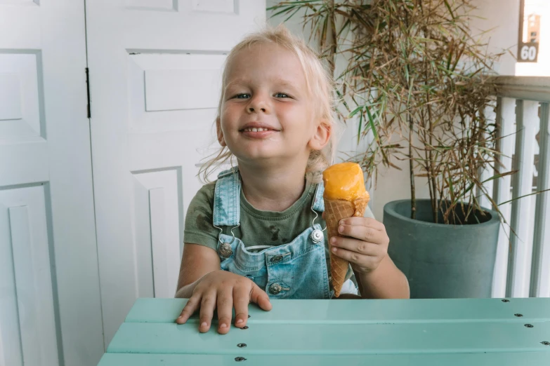 a little girl sitting at a table eating an ice cream cone, pexels contest winner, holding a tangerine, blond boy, blue overalls, she is smiling