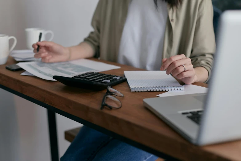 a woman sitting at a table with a laptop and a calculator, trending on pexels, three quater notes, low quality photo, thumbnail, holding notebook