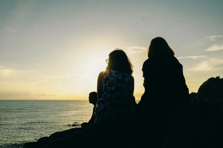 a couple of women sitting on top of a rock next to the ocean, pexels, silhouette :7, profile image, good friends, dark warm light