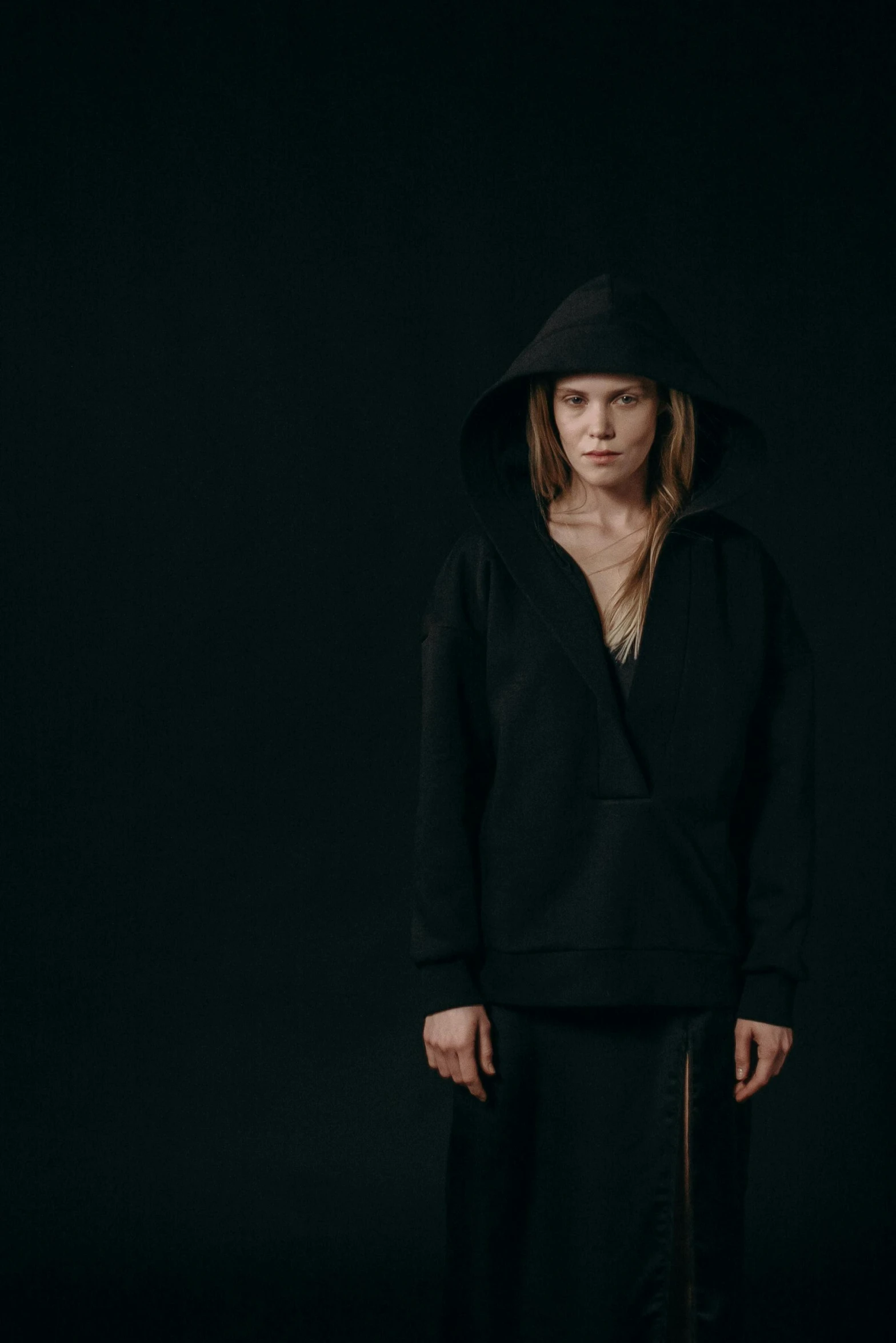 a woman standing in front of a black background, inspired by Louisa Matthíasdóttir, minimalism, hat and hoodie, concerned, press shot, in style of britt marling