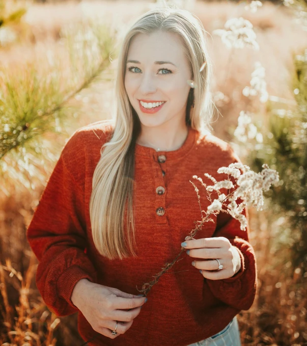 a woman standing in a field holding a flower, by Kristin Nelson, pexels contest winner, light stubble with red shirt, brown sweater, portrait of maci holloway, casey cooke