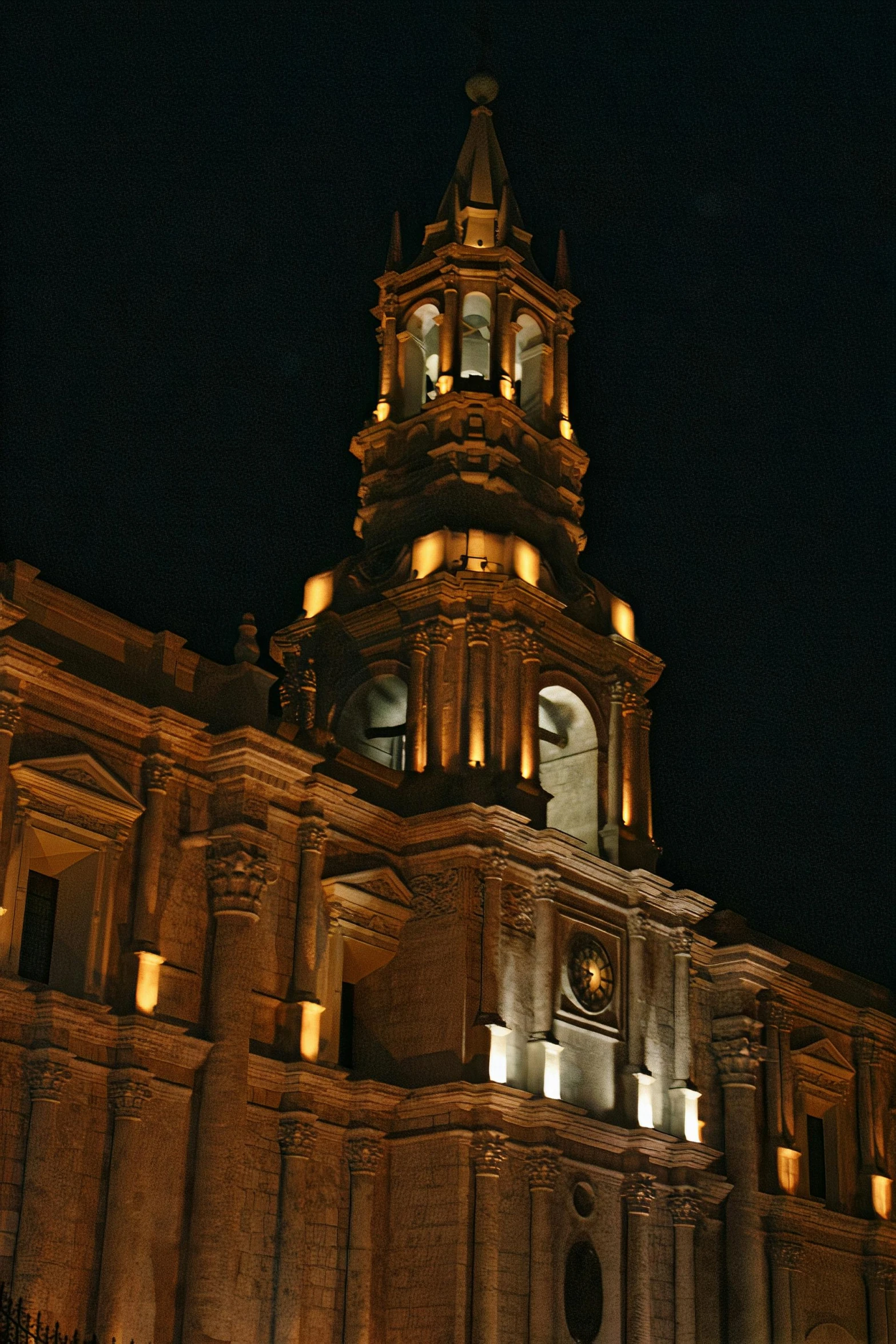 a large building with a clock tower lit up at night, inspired by Francisco Zúñiga, baroque architecture, neo - andean architecture, dramatic lighting - n 9, spire