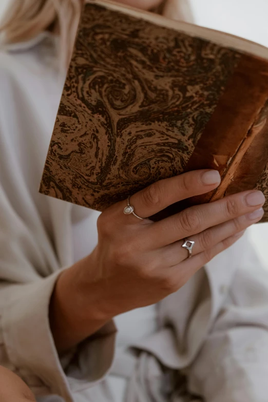 a woman sitting on a couch reading a book, an album cover, by Alice Mason, trending on unsplash, renaissance, wearing two metallic rings, close-up of thin soft hand, nordic wedding ring, handcrafted paper background