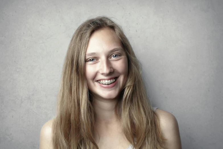 a beautiful young woman standing in front of a gray wall, by Matthias Stom, pexels contest winner, renaissance, cute smiling face, hyperrealistic teen, brown-blond-hair pretty face, scientific photo