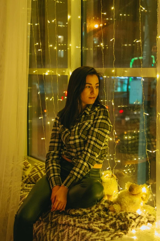 a woman sitting on top of a bed next to a window, a picture, inspired by Elsa Bleda, pexels contest winner, hurufiyya, wearing festive clothing, yellow lights, wearing plaid shirt, 🤤 girl portrait