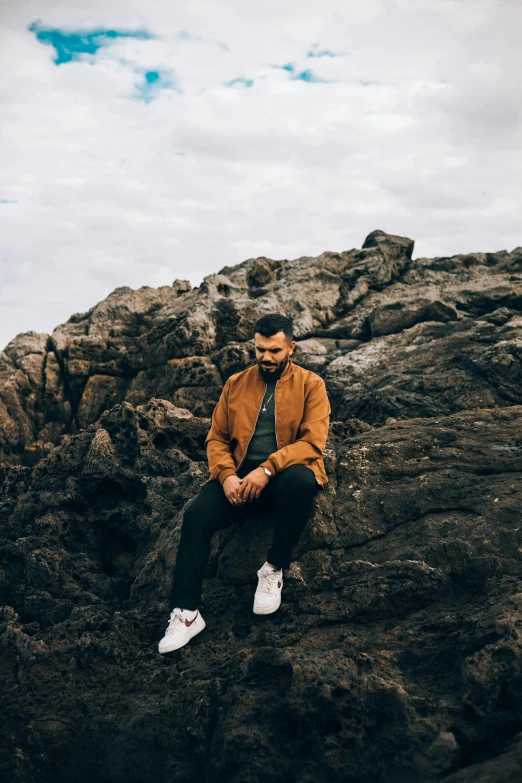 a man sitting on a rock near the ocean, an album cover, pexels contest winner, wearing a bomber jacket, a portrait of rahul kohli, caramel. rugged, tessellated planes of rock