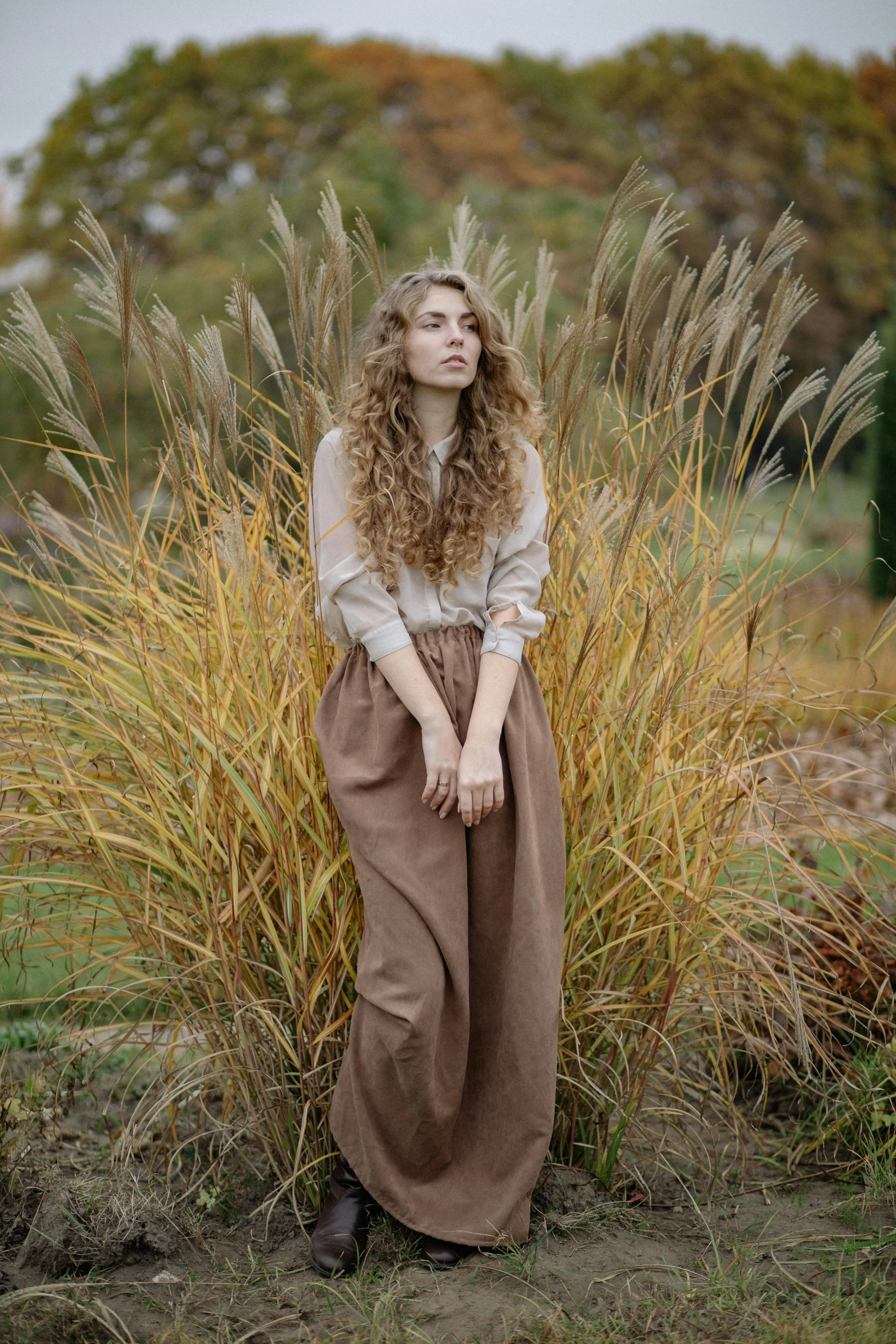 a woman standing in front of some tall grass, by irakli nadar, renaissance, brown pants, curls, in autumn, portrait image