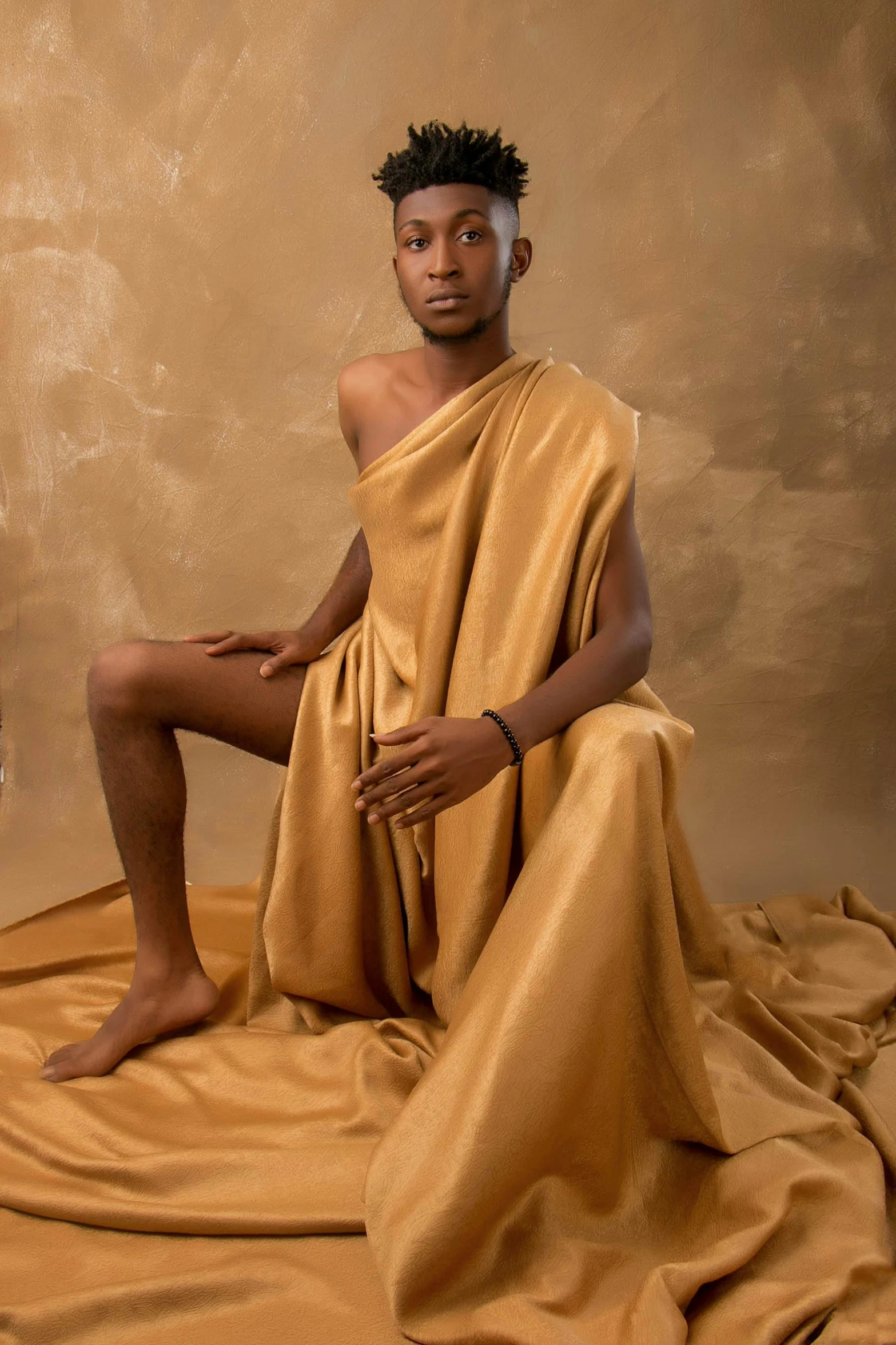 a man sitting on top of a bed covered in a blanket, an album cover, inspired by Thomas Blackshear, trending on pexels, hyperrealism, golden dress, yoruba body paint, shaven, at a fashion shoot