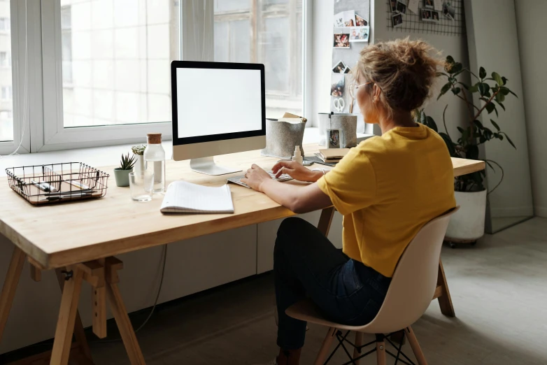 a woman sitting at a desk in front of a computer, trending on pexels, wearing a modern yellow tshirt, focus her back, profile image, scandinavian design