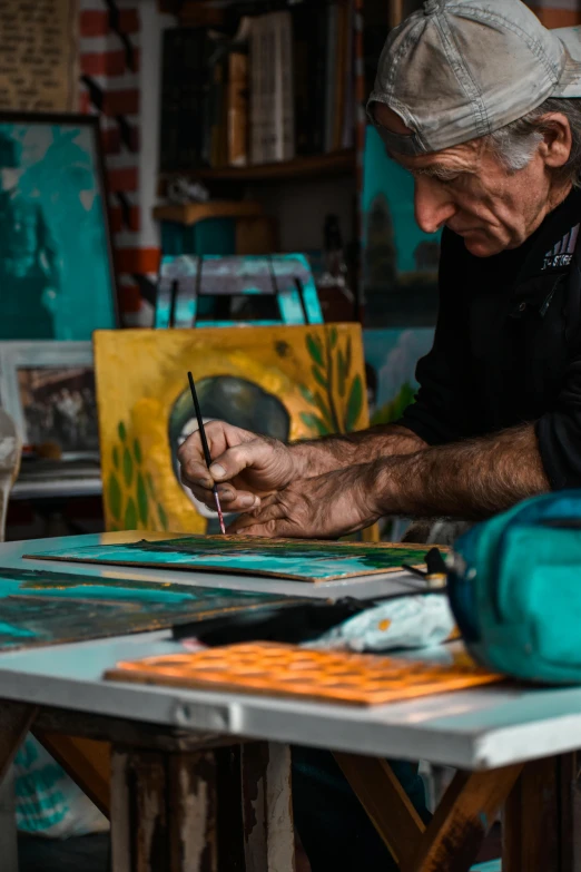 a man sitting at a table working on a painting, trending on pexels, arbeitsrat für kunst, teal and orange colors, old man doing hard work, center of image, painting on black canvas