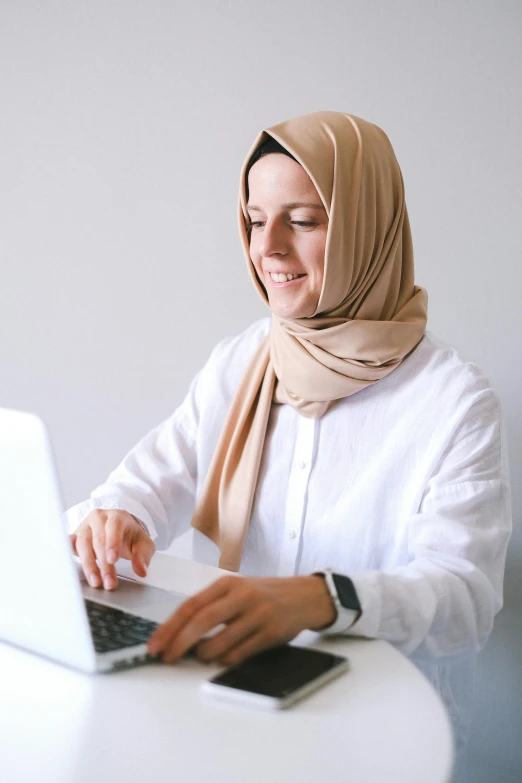 a woman sitting at a table using a laptop computer, inspired by Maryam Hashemi, pexels contest winner, hurufiyya, white scarf, wearing business casual dress, beige, caucasian