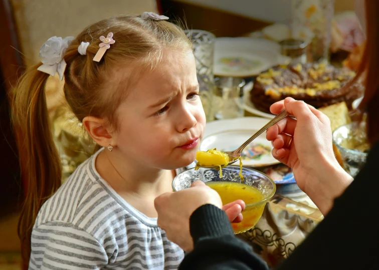 a little girl sitting at a table with a spoon in her mouth, by Adam Marczyński, pexels, renaissance, disgusted, good soup, square, celebration