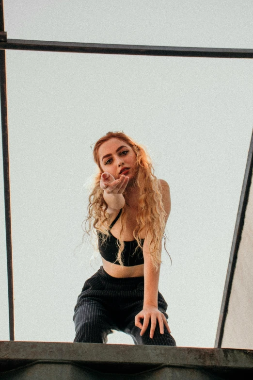 a woman standing on a ledge talking on a cell phone, an album cover, inspired by Elsa Bleda, trending on pexels, wearing a cropped black tank top, sitting pose, curly blond, sky - high view