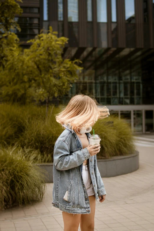 a woman standing on a sidewalk with her hair blowing in the wind, pexels contest winner, iced latte, jean jacket, walking at the garden, sunfaded