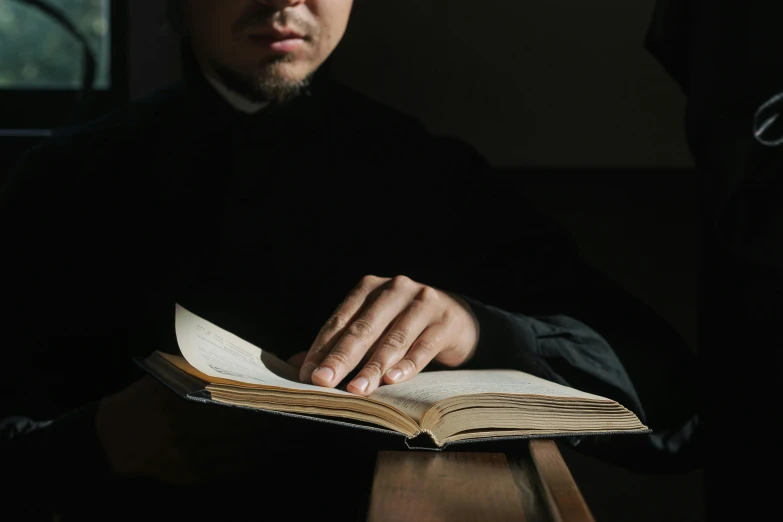 a man sitting at a table reading a book, unsplash, hurufiyya, an evil catholic priest, asian male, woman in black robes, istock
