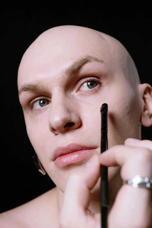 a close up of a person with a bald head, an album cover, inspired by Seb McKinnon, featured on reddit, antipodeans, putting makeup on, holding pencil, vitaly bugarov, nonbinary model