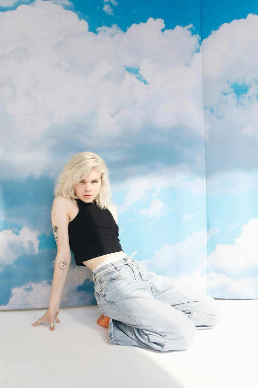 a woman sitting on top of a white floor next to a wall, an album cover, inspired by Elsa Bleda, trending on pexels, tattoos of cumulus clouds, portrait anya taylor-joy, jeans, on clouds